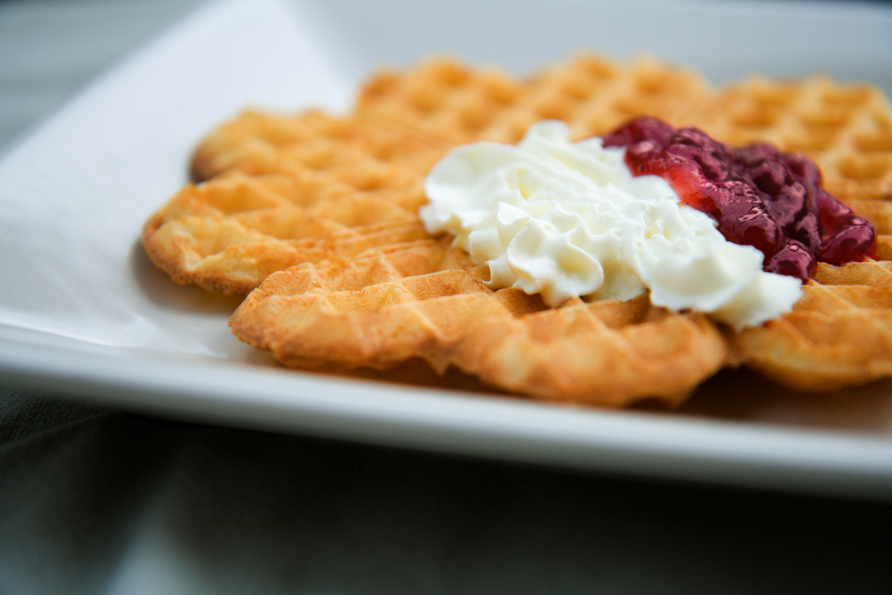 A close-up of a fresh swedish style waffle on a sqaure plate. Strawberry jam and whipped cream on top of the waffle. Shallow dept of field.